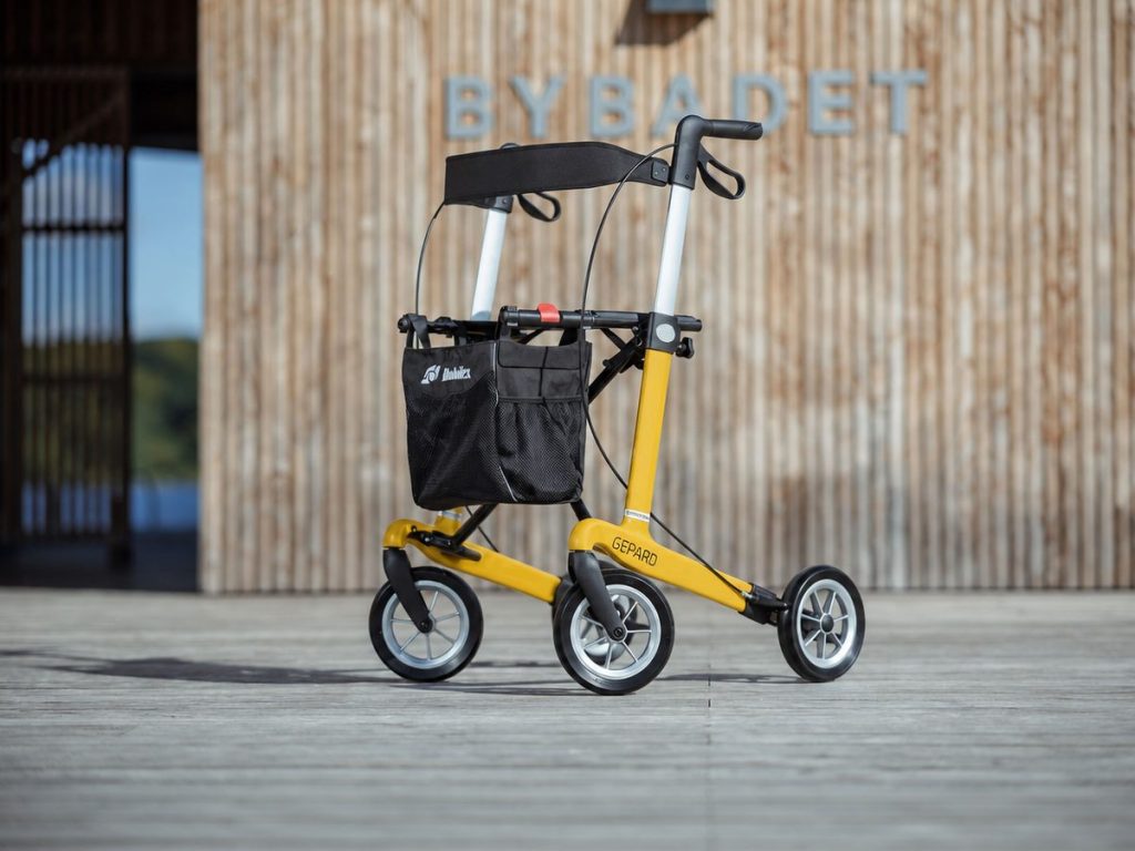 Carbon-Rollator Gepard in Gelb mit Einkaufstasche und Rückengurt vor einer Holzfassade